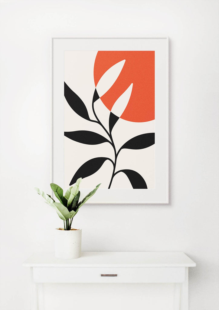 Floral Abstract Wall Art by Wall Funk features black leaves on an orange circle, framed above a plant on a white table.