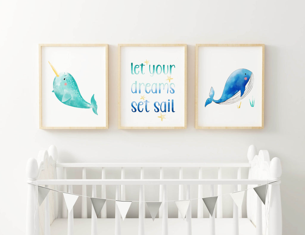 Three framed Wall Funk prints above crib: narwhal, "let your dreams set sail," whale. Silver banner on the crib.